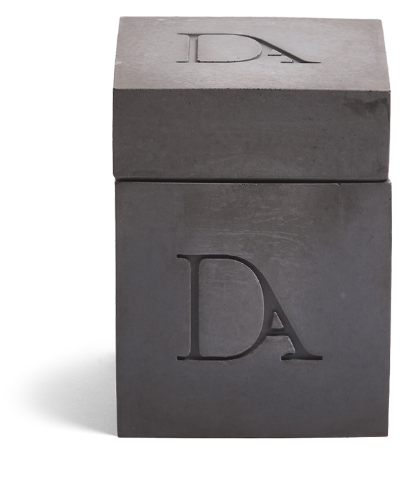9oz Devetu artisanal candle in Apollo, encased in gunmetal black concrete square vessel with a lid. The lid, featuring the embossed 'DA' Devetu Aroma logo, adds a sleek finish to the candle's modern aesthetic.