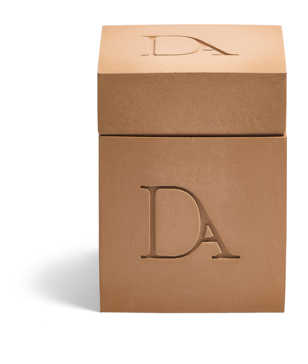 9oz Devetu artisanal candle in Embrace, encased in warm cashew colored concrete square vessel with a lid. The lid, featuring the embossed 'DA' Devetu Aroma logo, adds a sleek finish to the candle's modern aesthetic.