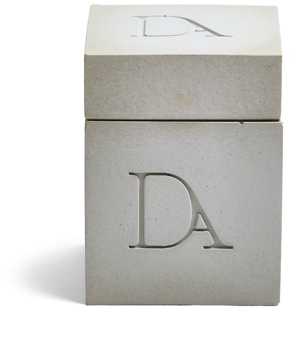 9oz Devetu artisanal candle in Skin, encased in a natural concrete square vessel with a lid. The lid, featuring the embossed 'DA' Devetu Aroma logo, complements the minimalist aesthetic.