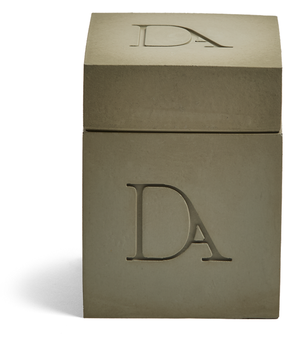 9oz Devetu artisanal candle in Wild Nomad, encased in a military green concrete square vessel with a lid. The lid, featuring the embossed 'DA' Devetu Aroma logo, enhances the candle's robust and earthy charm.