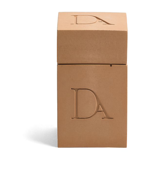 4oz Devetu artisanal candle in Embrace, encased in warm cashew colored concrete square vessel with a lid. The lid, featuring the embossed 'DA' Devetu Aroma logo, adds a sleek finish to the candle's modern aesthetic.