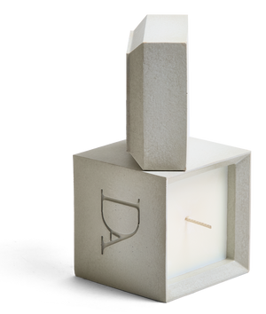 9oz Devetu artisanal candle in Skin, encased in a natural concrete square vessel with a lid. The lid, featuring the embossed 'DA' Devetu Aroma logo, complements the minimalist aesthetic.