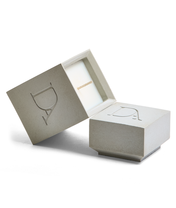 4oz Devetu artisanal candle in Skin, encased in natural concrete gray concrete square vessel with a lid. The lid, featuring the embossed 'DA' Devetu Aroma logo, adds a sleek finish to the candle's modern aesthetic.