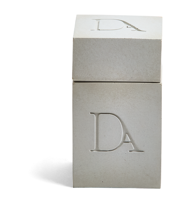 4oz Devetu artisanal candle in Skin, encased in natural concrete gray concrete square vessel with a lid. The lid, featuring the embossed 'DA' Devetu Aroma logo, adds a sleek finish to the candle's modern aesthetic.